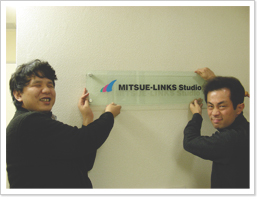 写真：ミツエーリンクス スタジオの看板に手を掛ける辻ちゃん・ウエちゃん